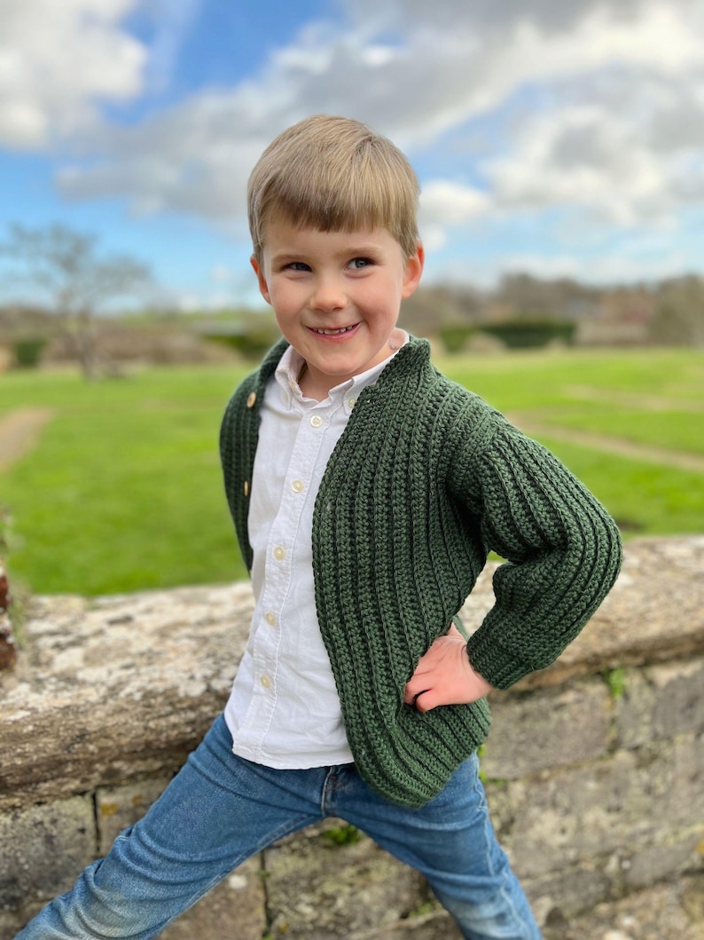 K&ouml;nnte beinhalten: Ein kleiner Junge tr&auml;gt einen dunkelgr&uuml;nen Strickcardigan mit einem wei&szlig;en Hemd mit Kn&ouml;pfen und blauen Jeans. Er steht vor einer Steinmauer mit einem grasbewachsenen Feld im Hintergrund.