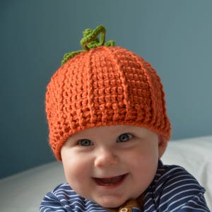 May include: A baby wearing a crocheted pumpkin hat with a green stem. The hat is orange and the baby is smiling.