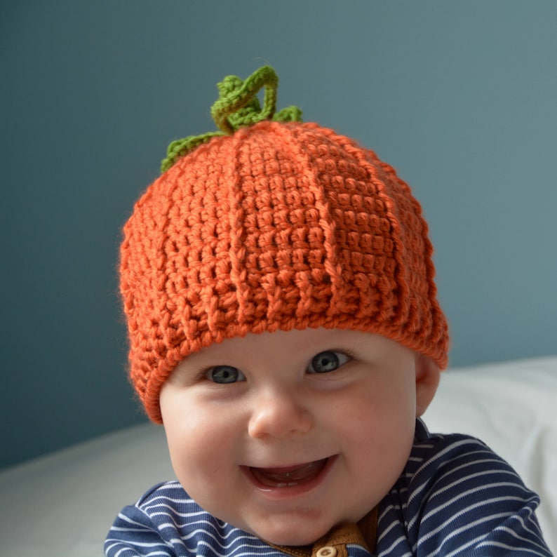 May include: A baby wearing a crocheted pumpkin hat with a green stem. The hat is orange and the baby is smiling.