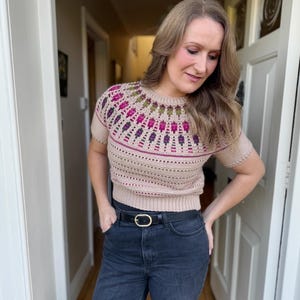 Pu&ograve; includere: Un maglione beige a maniche corte con un design a giogo colorato e dettagliato. Il giogo presenta un motivo di forme rosa, viola e verdi. Il maglione &egrave; abbinato a jeans neri e una cintura nera con fibbia dorata.