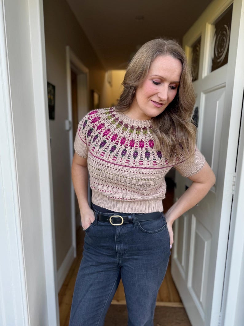 Pu&ograve; includere: Un maglione beige a maniche corte con un design a giogo colorato e dettagliato. Il giogo presenta un motivo di forme rosa, viola e verdi. Il maglione &egrave; abbinato a jeans neri e una cintura nera con fibbia dorata.