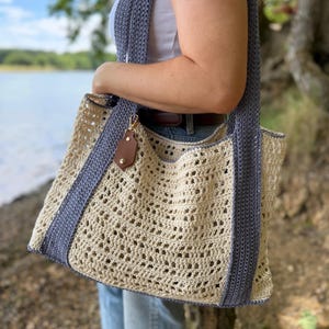 Puede incluir: Un bolso tote de ganchillo con cuerpo calado de color crema y correas y bordes de color azul b&iacute;garo. El bolso incluye una peque&ntilde;a etiqueta de cuero sint&eacute;tico marr&oacute;n sujeta a un cierre de metal dorado cerca del borde superior del compartimento.