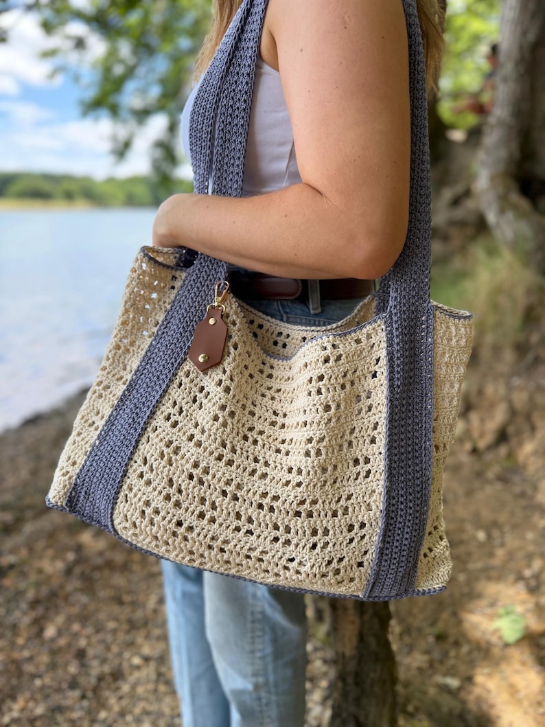 Puede incluir: Un bolso tote de ganchillo con cuerpo calado de color crema y correas y bordes de color azul b&iacute;garo. El bolso incluye una peque&ntilde;a etiqueta de cuero sint&eacute;tico marr&oacute;n sujeta a un cierre de metal dorado cerca del borde superior del compartimento.