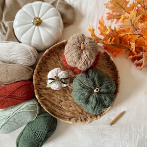 May include: An autumnal still life featuring decorative pumpkins and yarn. A white ceramic pumpkin with a gold stem is next to a woven basket with knitted pumpkins in brown and green. Skeins of yarn and autumn leaves complete the display.