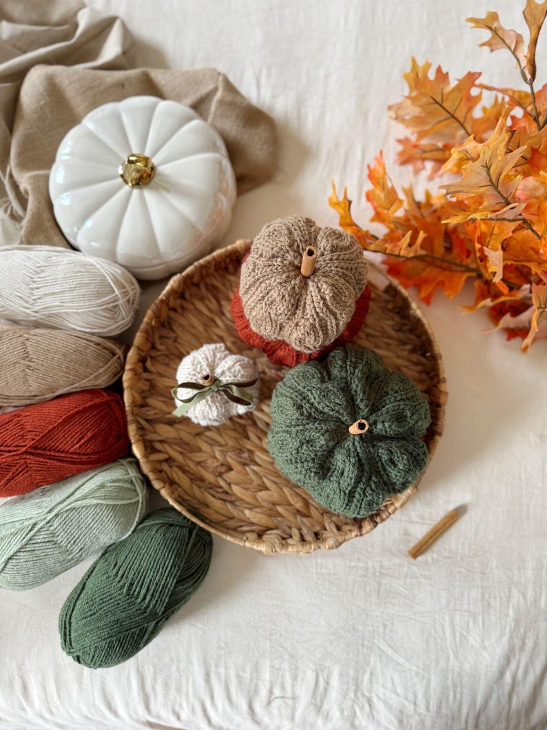 May include: An autumnal still life featuring decorative pumpkins and yarn. A white ceramic pumpkin with a gold stem is next to a woven basket with knitted pumpkins in brown and green. Skeins of yarn and autumn leaves complete the display.