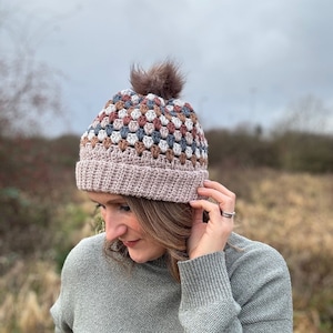 May include: A crocheted beanie with a pom pom in shades of brown, grey, and white. The beanie has a textured stitch pattern and a solid pink brim.