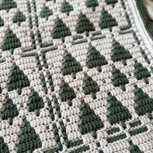 May include: A crochet blanket featuring a repeating geometric pattern of dark forest green pine trees against a cream-colored background. The textile displays a textured stitch construction with a thick, solid dark green border along the edge.