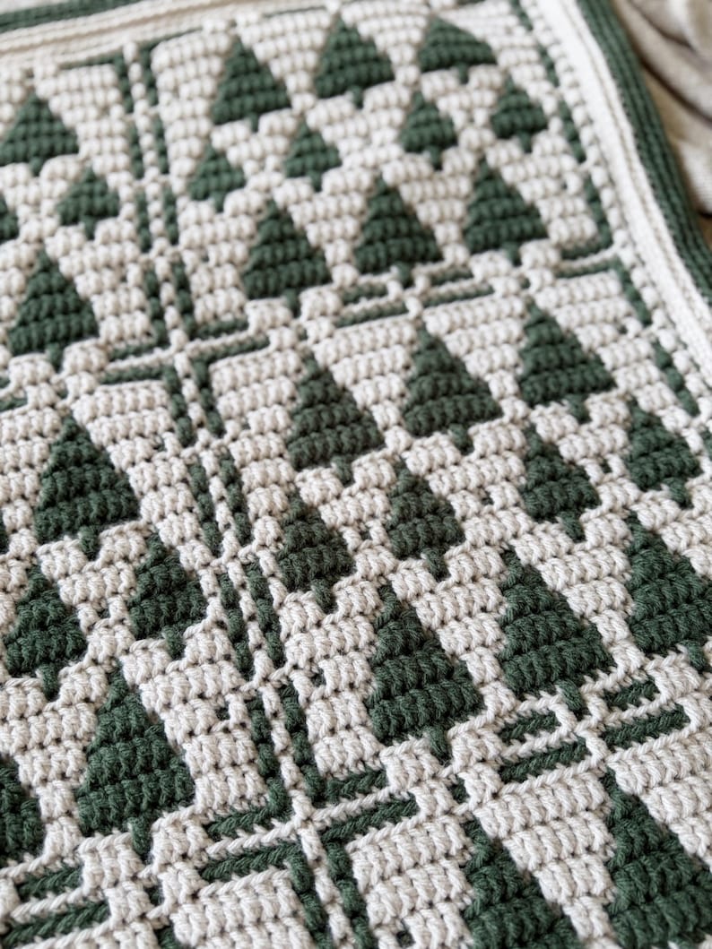 May include: A crochet blanket featuring a repeating geometric pattern of dark forest green pine trees against a cream-colored background. The textile displays a textured stitch construction with a thick, solid dark green border along the edge.