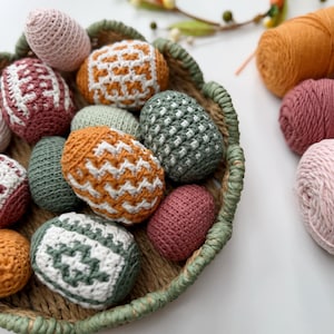 May include: A woven basket filled with various crocheted Easter eggs in shades of pink, green, orange, and white. Some eggs have patterned designs. A wreath and yarn skeins are in the background.