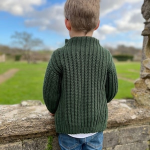 K&ouml;nnte beinhalten: Eine junge Person tr&auml;gt einen dunkelgr&uuml;nen Strickpullover mit Stehkragen und blaue Jeans. Die Person steht vor einer Steinmauer und blickt auf ein grasbewachsenes Feld.