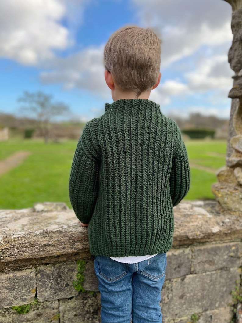 K&ouml;nnte beinhalten: Eine junge Person tr&auml;gt einen dunkelgr&uuml;nen Strickpullover mit Stehkragen und blaue Jeans. Die Person steht vor einer Steinmauer und blickt auf ein grasbewachsenes Feld.