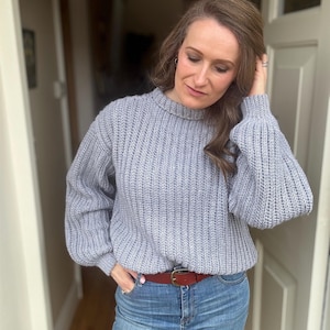 May include: A pale grey ribbed knit jumper with a crew neck and long, balloon sleeves. The jumper is worn with blue jeans and a brown leather belt with a gold buckle. The outfit is casual and comfortable.