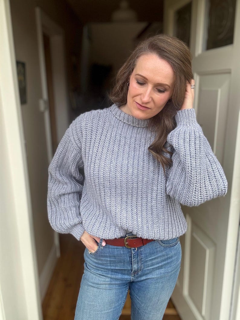 May include: A pale grey ribbed knit jumper with a crew neck and long, balloon sleeves. The jumper is worn with blue jeans and a brown leather belt with a gold buckle. The outfit is casual and comfortable.
