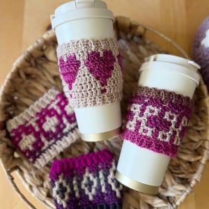 Op de afbeelding: Een verzameling handgemaakte gehaakte bekerhoezen in een gevlochten mand. De hoezen hebben verschillende geometrische patronen en hartpatronen in roze, paars en beige tinten. Twee hoezen zitten op witte reisbekers.