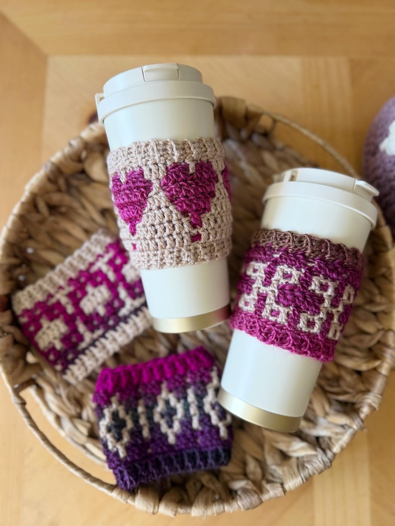 Op de afbeelding: Een verzameling handgemaakte gehaakte bekerhoezen in een gevlochten mand. De hoezen hebben verschillende geometrische patronen en hartpatronen in roze, paars en beige tinten. Twee hoezen zitten op witte reisbekers.