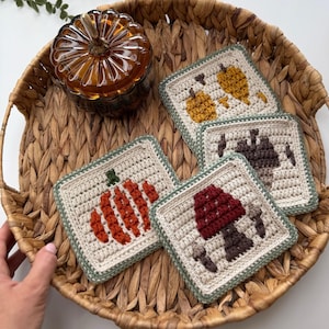 May include: Four square crochet coasters featuring autumn-themed designs in cream yarn with sage green borders. The designs include a bright orange pumpkin, a red and brown mushroom, a brown acorn, and a yellow leaf, all worked in textured stitches.