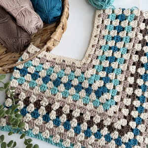 May include: A crocheted garment with a v-neck, made with beige, brown, teal, and light blue yarn. The garment is laid flat on a white surface with balls of yarn in the background.