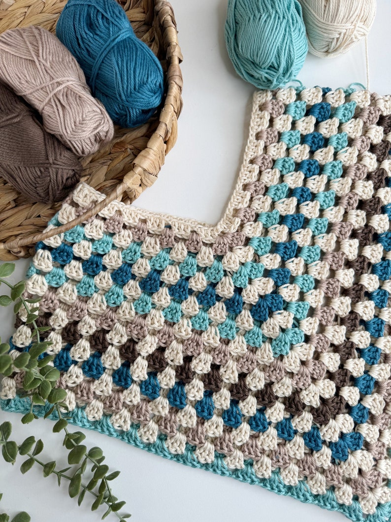 May include: A crocheted garment with a v-neck, made with beige, brown, teal, and light blue yarn. The garment is laid flat on a white surface with balls of yarn in the background.
