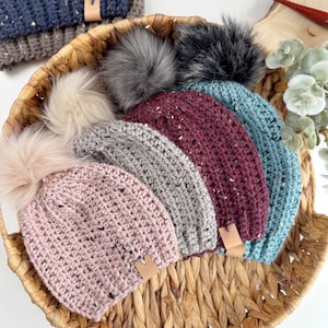 Puede incluir: Cinco gorros de punto con pompones de piel sintética en una cesta de mimbre. Los gorros son de color rosa, gris, burdeos y azul.