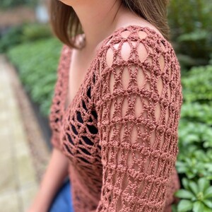 Puede incluir: Un top de crochet de color terracota con un dise&ntilde;o suelto y tejido abierto. El top tiene un cuello en V y mangas tres cuartos.