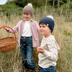 May include: Two children wearing knit beanies with faux fur pom-poms. One beanie is brown, the other is navy blue. One child wears a pink shirt and carries a wicker basket. The other child wears a striped shirt.