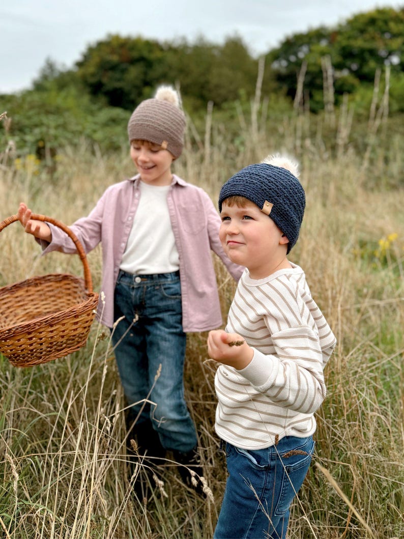 May include: Two children wearing knit beanies with faux fur pom-poms. One beanie is brown, the other is navy blue. One child wears a pink shirt and carries a wicker basket. The other child wears a striped shirt.