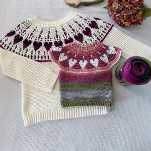 May include: A cream-colored long-sleeve sweater with a yoke featuring a dark purple and green heart pattern, paired with a smaller matching short-sleeve sweater in a gradient of pink, purple, green, and gray, accompanied by a ball of variegated yarn.