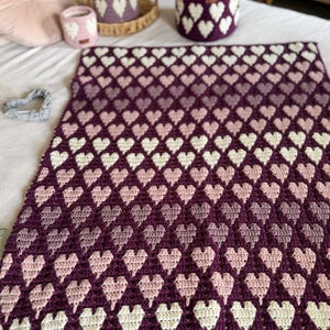 Op de afbeelding: Een handgemaakte gehaakte deken met een hartpatroon in tinten paars, roze en wit. De afbeelding toont ook bijpassende gehaakte manden, waardoor een geco&ouml;rdineerde woondecoratieset ontstaat. De afmetingen van de deken zijn niet zichtbaar.