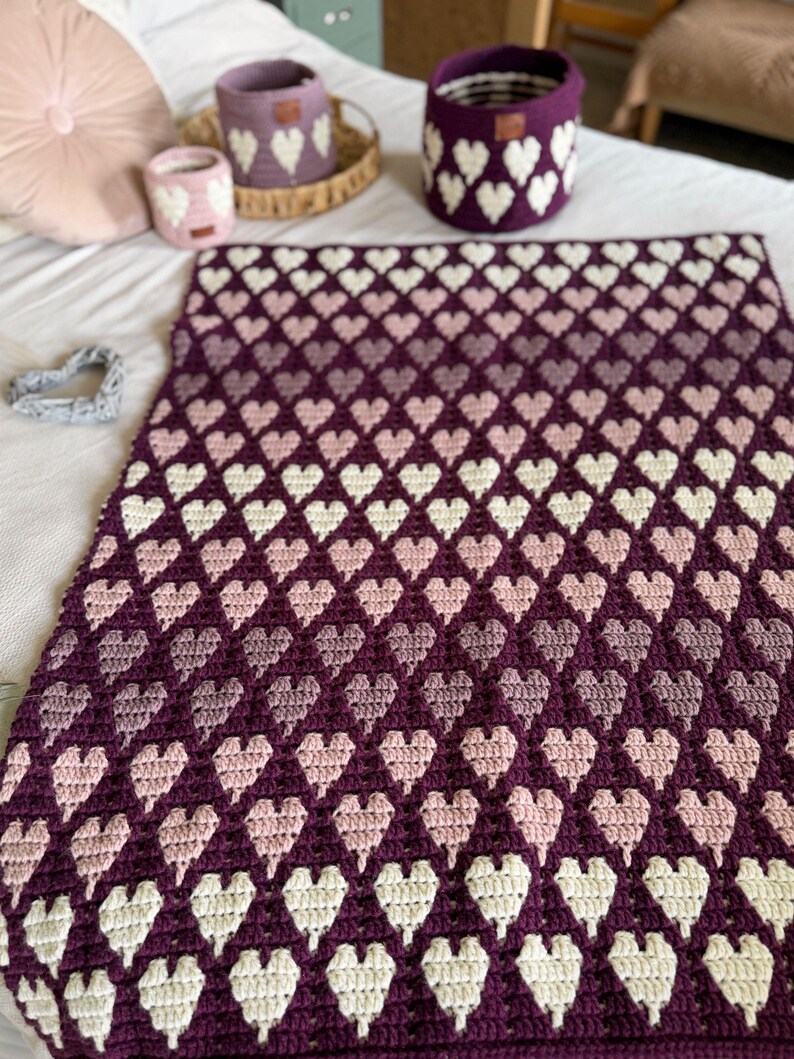 Op de afbeelding: Een handgemaakte gehaakte deken met een hartpatroon in tinten paars, roze en wit. De afbeelding toont ook bijpassende gehaakte manden, waardoor een geco&ouml;rdineerde woondecoratieset ontstaat. De afmetingen van de deken zijn niet zichtbaar.