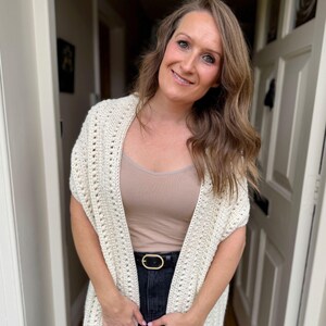 May include: A cream-colored, open-front crochet shawl with a textured pattern. The shawl is worn over a neutral-colored top and black jeans, accessorized with a black belt with a gold buckle. The outfit is casual and comfortable.