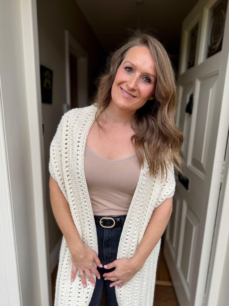 May include: A cream-colored, open-front crochet shawl with a textured pattern. The shawl is worn over a neutral-colored top and black jeans, accessorized with a black belt with a gold buckle. The outfit is casual and comfortable.
