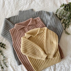 May include: Three knitted jumpers in grey, blush pink, and pale yellow. The grey jumper has a ribbed turtleneck, while the pink and yellow jumpers have collared necklines. All jumpers have long sleeves and are made of a thick, textured yarn.