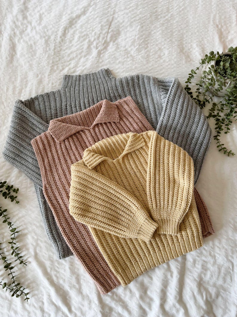 May include: Three knitted jumpers in grey, blush pink, and pale yellow. The grey jumper has a ribbed turtleneck, while the pink and yellow jumpers have collared necklines. All jumpers have long sleeves and are made of a thick, textured yarn.
