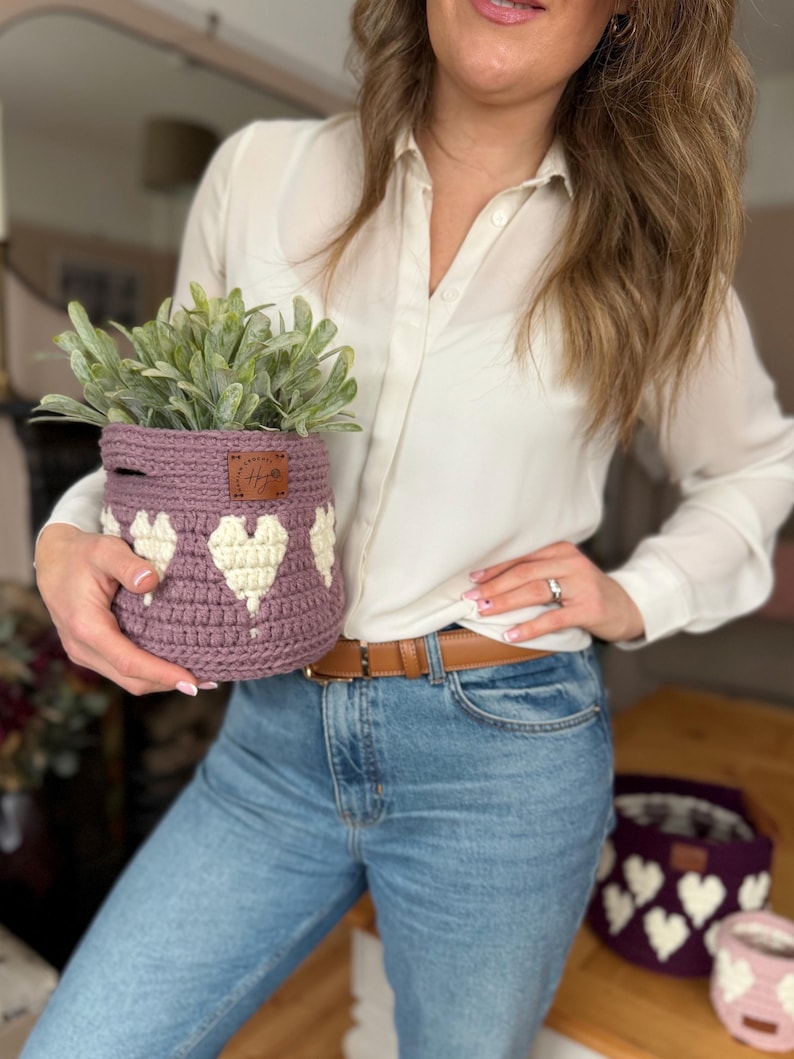 Op de afbeelding: Een gehaakte lavendelkleurige mand met witte hartpatronen, met een groene plant. De mand heeft een bruin leren label. Een andere mand is zichtbaar op de achtergrond.