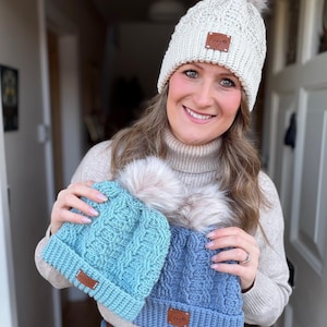 May include: A woman holding three knitted beanies with faux fur pom-poms. One is cream-colored, and the others are light blue and blue. Each beanie has a brown leather tag. The beanies have a cable knit design.