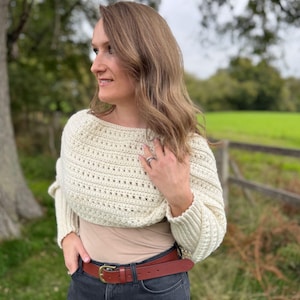May include: Cream-colored crochet shrug with long sleeves. The shrug is worn over a neutral-colored top and paired with a brown leather belt with a gold buckle and dark wash jeans. The background features a green field and trees.