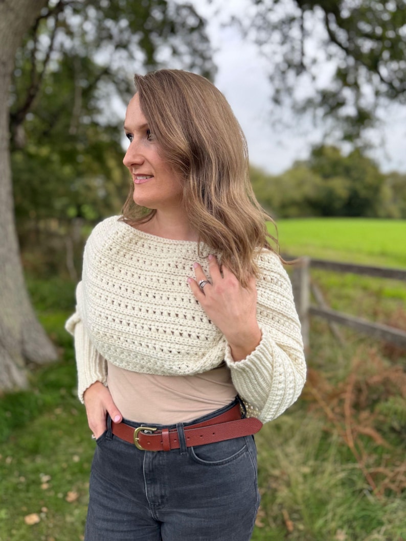 May include: Cream-colored crochet shrug with long sleeves. The shrug is worn over a neutral-colored top and paired with a brown leather belt with a gold buckle and dark wash jeans. The background features a green field and trees.