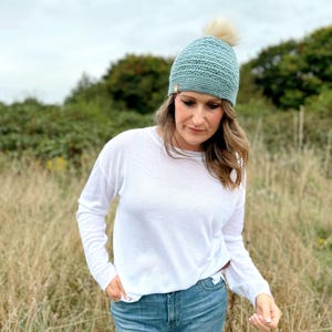 May include: A light blue crocheted beanie with a cream-colored pom-pom. The hat is worn by a person wearing a white long-sleeve shirt and blue jeans. The background features tall grass and green trees.