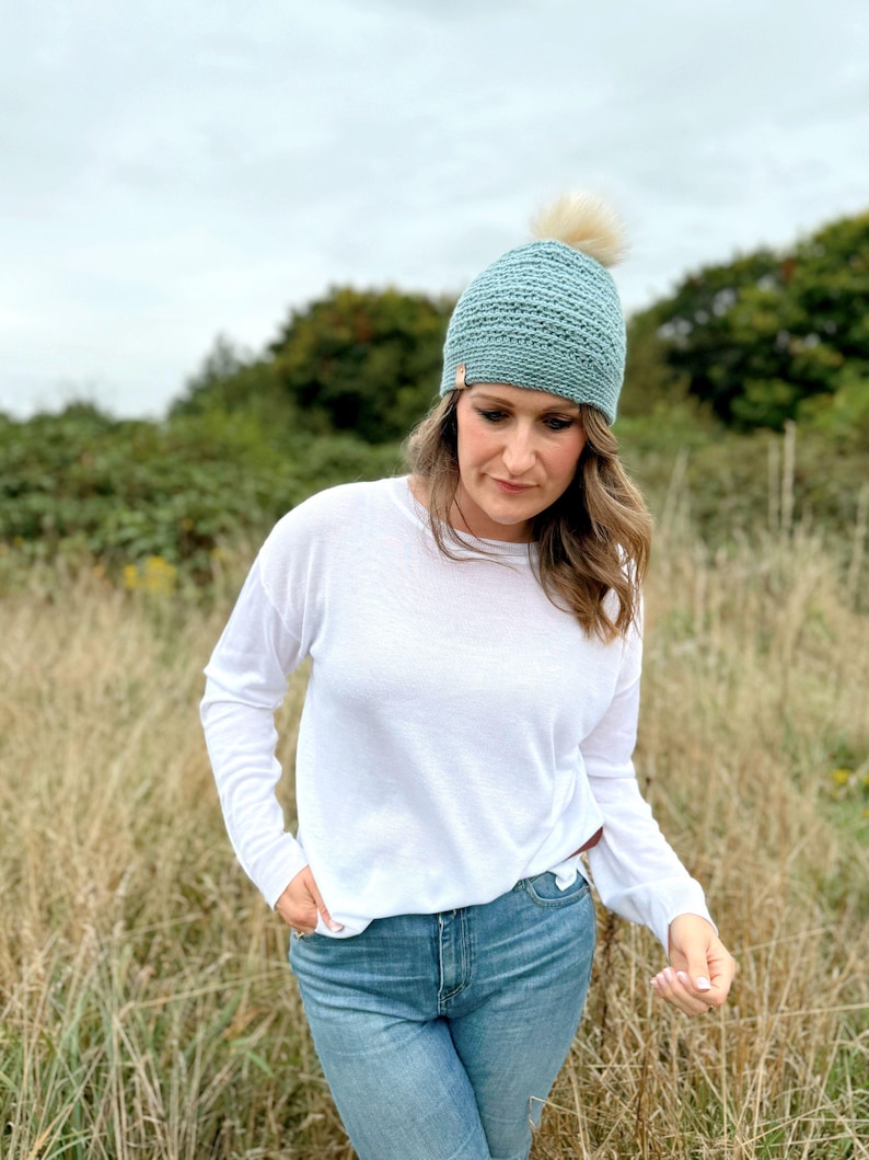 May include: A light blue crocheted beanie with a cream-colored pom-pom. The hat is worn by a person wearing a white long-sleeve shirt and blue jeans. The background features tall grass and green trees.