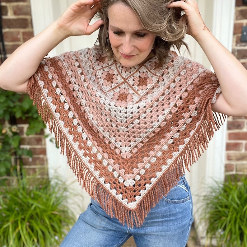 Puede incluir: Un poncho de ganchillo con un patr&oacute;n geom&eacute;trico en tonos marrones, rosas y blancos. El poncho tiene un borde de flecos y lo lleva una persona con el pelo casta&ntilde;o.
