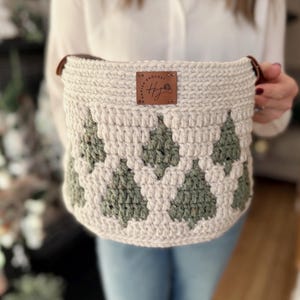 May include: A cream-coloured, crocheted basket with a green tree pattern. The basket has brown leather handles and a brown leather tag with the text "Hawaiian Crochet" and the initials "HJ".