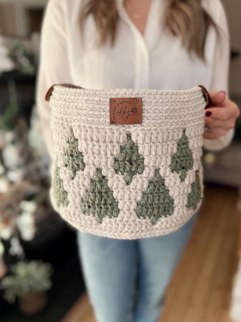 May include: A cream-coloured, crocheted basket with a green tree pattern. The basket has brown leather handles and a brown leather tag with the text "Hawaiian Crochet" and the initials "HJ".