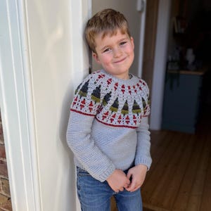 Könnte beinhalten: Hellblauer Häkelpullover mit detailliertem Passe, der ein Muster aus Bäumen und geometrischen Formen in Rot, Grün und Weiß aufweist. Der Pullover wird mit einer blauen Jeans kombiniert.