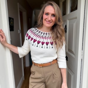 May include: A cream-colored, crocheted sweater with a pattern of burgundy hearts and black accents around the neckline. The sweater is paired with tan pleated trousers and a brown belt.