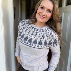 May include: A light beige sweater with a gray yoke featuring a geometric pattern and stylized tree designs. The long-sleeved sweater is made of a knitted material and has a ribbed hem and cuffs. The sweater is worn by a person.