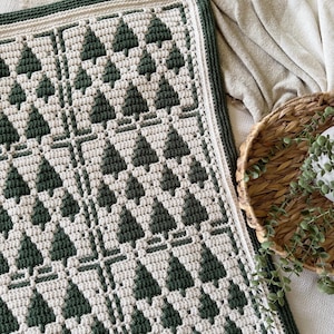 May include: A rectangular crochet blanket featuring a repeating pattern of dark forest green pine trees against a cream-colored background. The blanket is finished with a thick, dark green border and has a textured, woven appearance throughout.