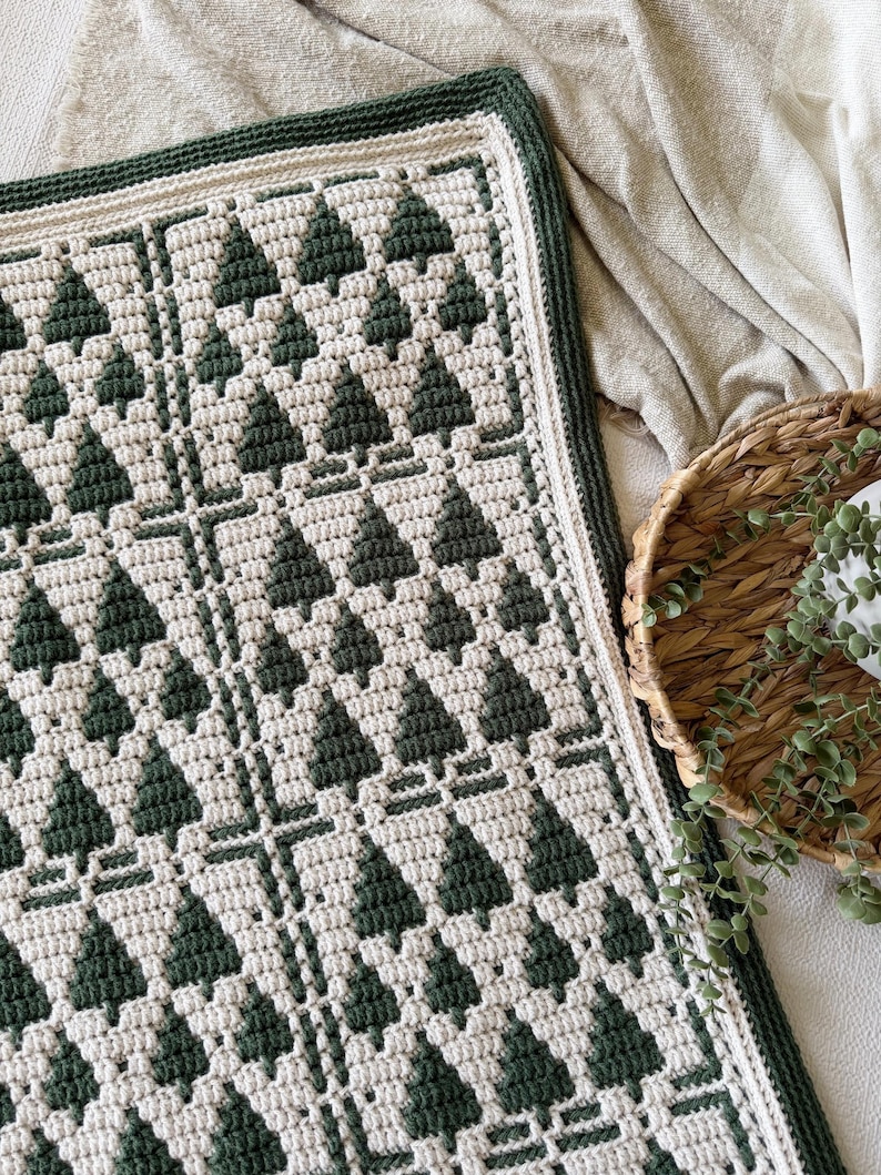 May include: A rectangular crochet blanket featuring a repeating pattern of dark forest green pine trees against a cream-colored background. The blanket is finished with a thick, dark green border and has a textured, woven appearance throughout.