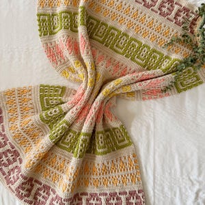 Könnte beinhalten: Eine handgehäkelte Decke mit einem geometrischen Muster in Beige, Grün, Rosa, Gelb und Lila. Die Decke ist über eine weiße Oberfläche drapiert und zeigt ihr kompliziertes Design und ihre strukturierten Maschen. Die Ränder der Decke weisen eine detaillierte Bordüre auf.