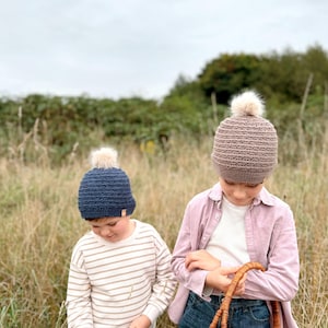 May include: Two children wearing knitted beanies with faux fur pom-poms. One beanie is navy blue, the other is taupe. One child wears a striped shirt, the other a pink shirt and jeans. They are in a field, one holding a wicker basket.
