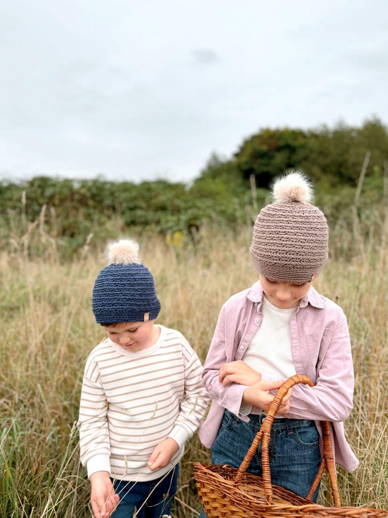 May include: Two children wearing knitted beanies with faux fur pom-poms. One beanie is navy blue, the other is taupe. One child wears a striped shirt, the other a pink shirt and jeans. They are in a field, one holding a wicker basket.